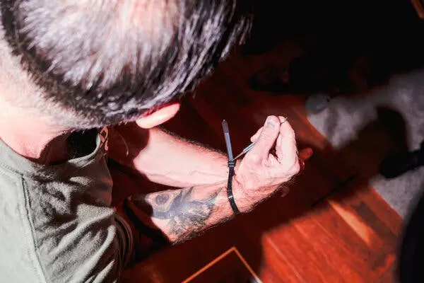 نگاهی از پشت شانهٔ فردی با تتوی بزرگ بر روی بازو که ابزار نازک فلزی‌اش را به داخل بند پلاستیکی سیاه که مچ‌هایش را به‌هم بسته، فشار می‌دهد.